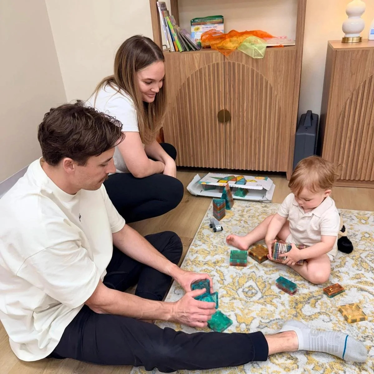 Two adults sit on the floor and build with magnetic tiles alongside a toddler